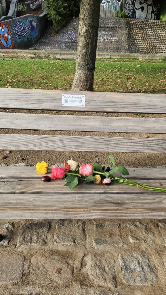Ausschnitt einer öffentlichen Bank mit einer Plakette, auf der "Ort des Gedenkens" steht. Auf der Bank liegen bunte Rosen und Kastanien. Im Hintergrund ist ein Baumstamm, eine Wiese und Gemäuer mit Graffiti