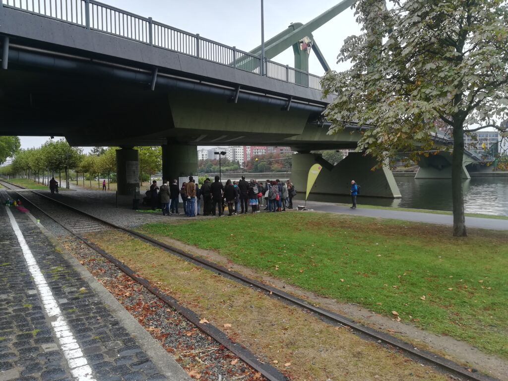 Eine Grasfläche am Main. Eine Gruppe Menschen steht für eine FRANS Veranstaltung unter einer Brücke.