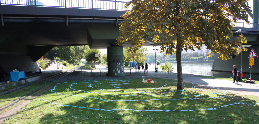 Grasfläche am Main mit Kastanie und einer Brücke im Hintergrund. Auf dem Gras liegen kreisförmig blaue Bänder. Menschen laufen auf dem Gehweg.
