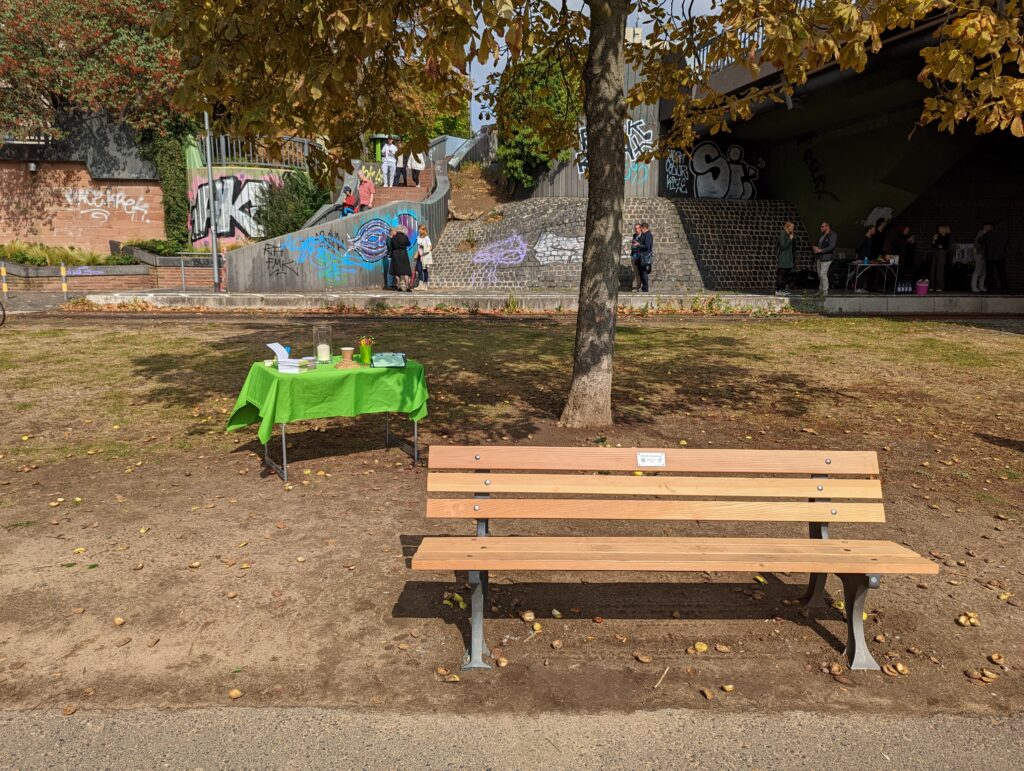 Eine ausgetrocknete Grasfläche am Main mit einer neu aufgestellten Bank. Im Hintergrund ein Infotisch mit grüner Decke, eine Kastanie und ein paar Menschen, die eine Treppe herunterlaufen oder unter einer Brücke stehen.