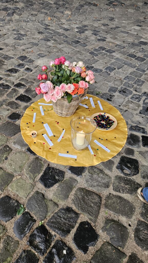 Kopfsteinpflaster am Boden und darauf liegt ein gelber kreisförmiger Stoff. Auf diesem steht eine brennende Kerze im Glas, bunte Rosen in einem Korb, buntes Kreis Konfetti sowie schmale Zettel mit Sprüchen.