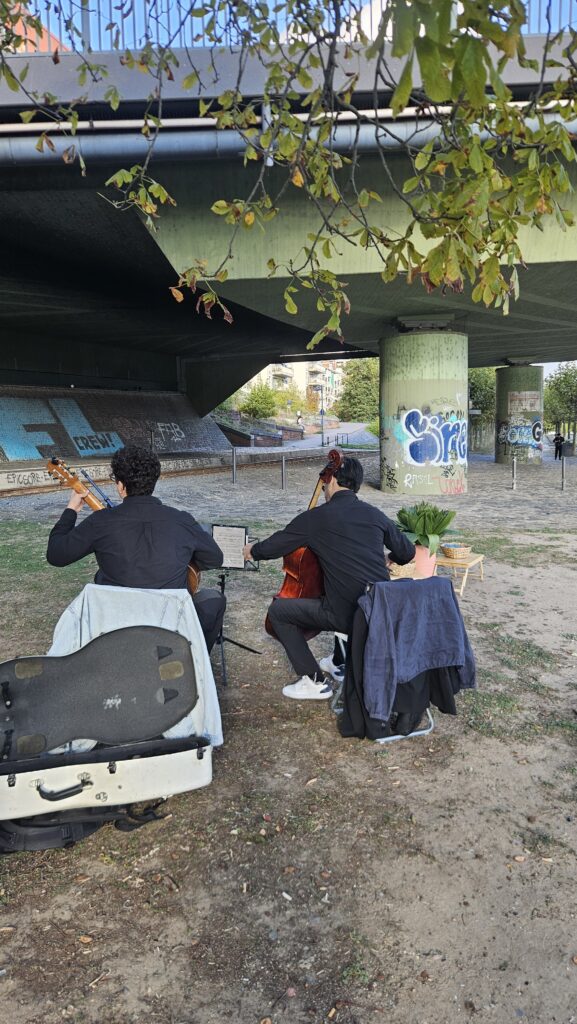 Blick auf eine Brücke, ein paar Äste einer Kastanie und zwei Musiker von hinten, die Gitarre und Cello spielen.