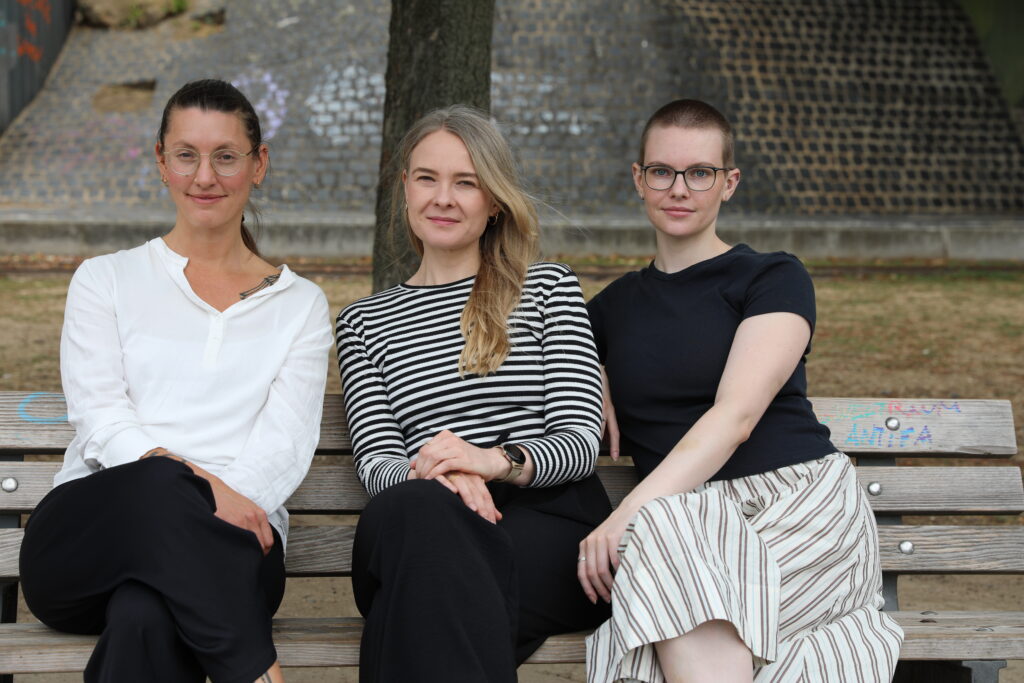 von links nach rechts: Nora Hauschild, Inga Beig und Victoria Dichter sitzen auf einer Bank und schauen in die Kamera