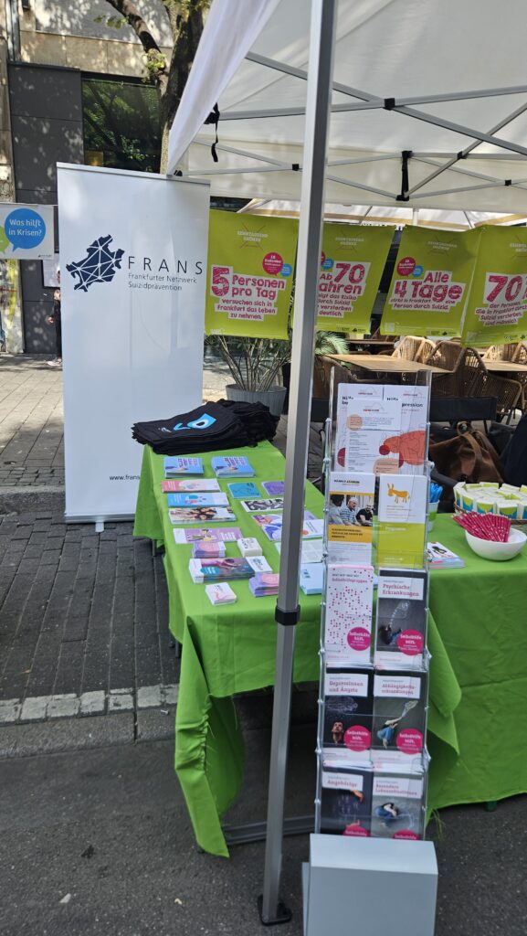 FRANS Infostand. Tische mit grünen Decken, einem weißen Roll-Up im Hintergrund, ebenso grüne Plakate mit Fakten zu Suizidalität. Auf den Tischen liegen Flyer und Give Aways. Vorne steht ein Aufsteller mit unterschiedlichen Flyern.