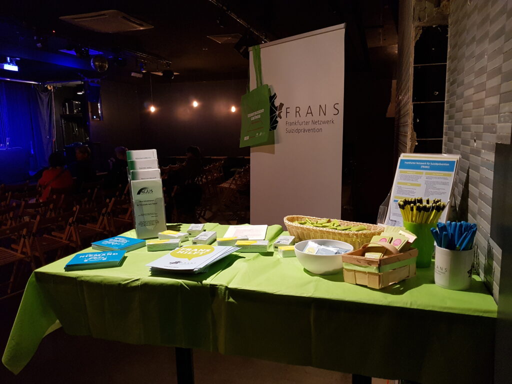 Dunkler Raum mit Theaterbestuhlung. Im Vordergrund des Bildes ein Tisch mit grüner Tischdecke und darauf viele Infomaterialen, Give Aways und im Hintergrund ein weißes FRANS Roll-Up an dessen Ecke ein grüner Beutel hängt.
