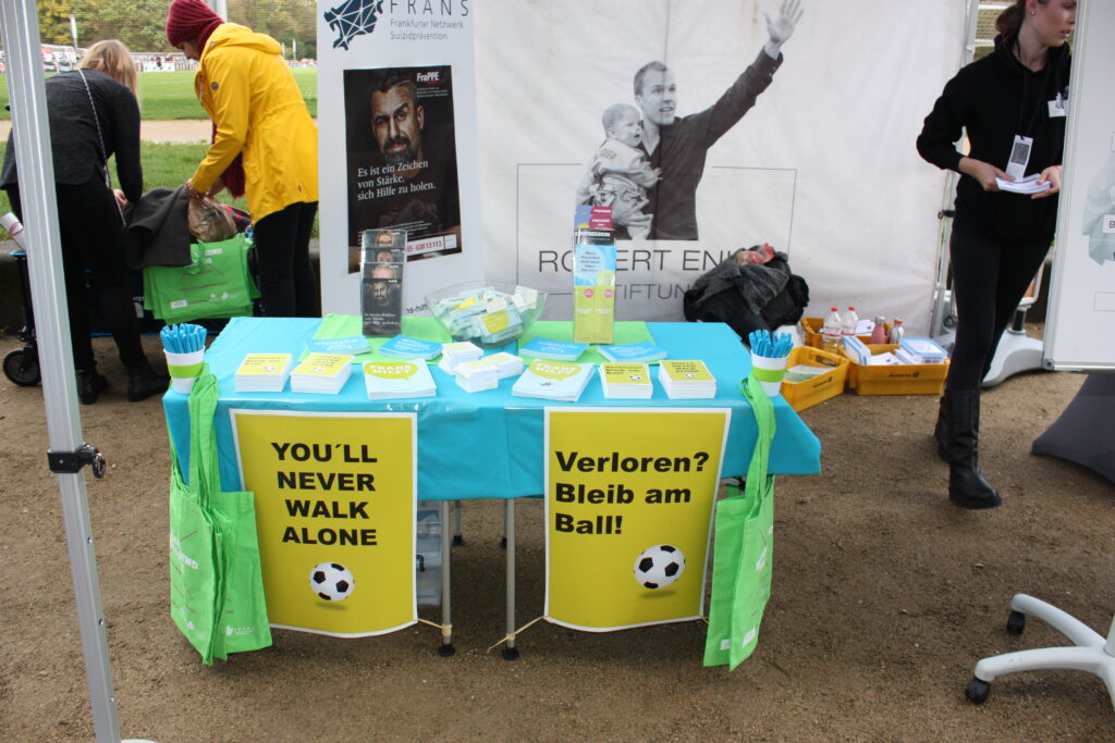 FRANS Infostand bei einem Fußballspiel. Tisch mit blauer Decke und darauf Infomaterial. An den Tischen hängen grüne Plakate mit der Aufschrift "You'll never walk alone" und "Verloren? Bleib am Ball!". Im Hintergrund sind Menschen, ein Roll-Up und ein Stoff-Plakat der Robert Enke Stiftung.