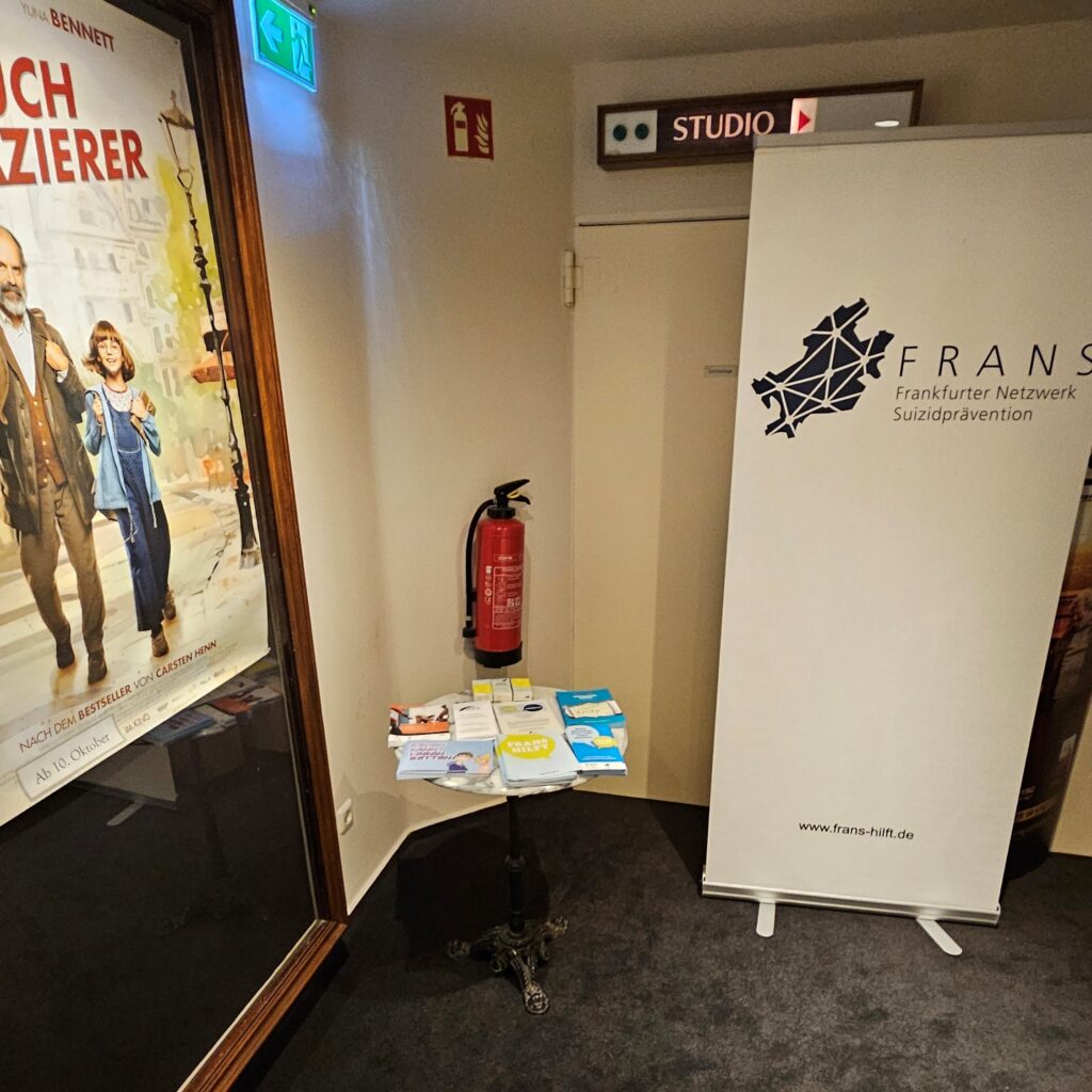 FRANS Infotisch vor einem Kinosaal. Links hängt ein Filmplakat und rechts im Bild steht ein weißes Roll-Up.