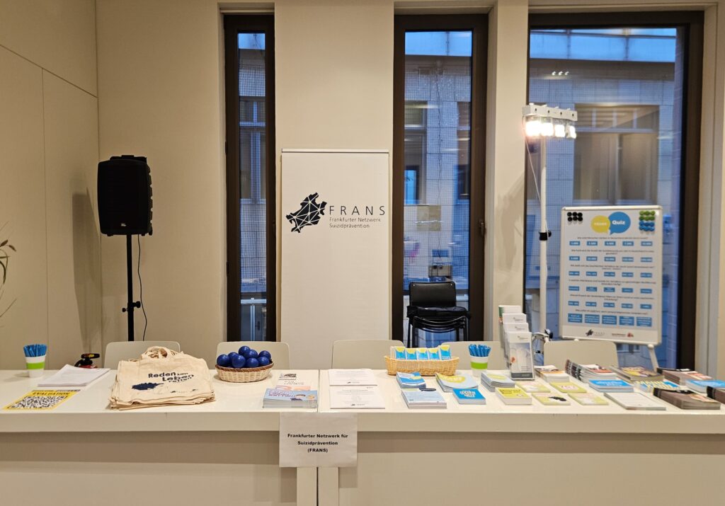Blick auf einen großen weißen Infotisch mit vielen Infomaterialien. Dahinter steht ein weißes Roll-Up und ein buntes FRANS Quiz. Dahinter sind große, tiefe Fenster mit Blick auf ein Nebengebäude.