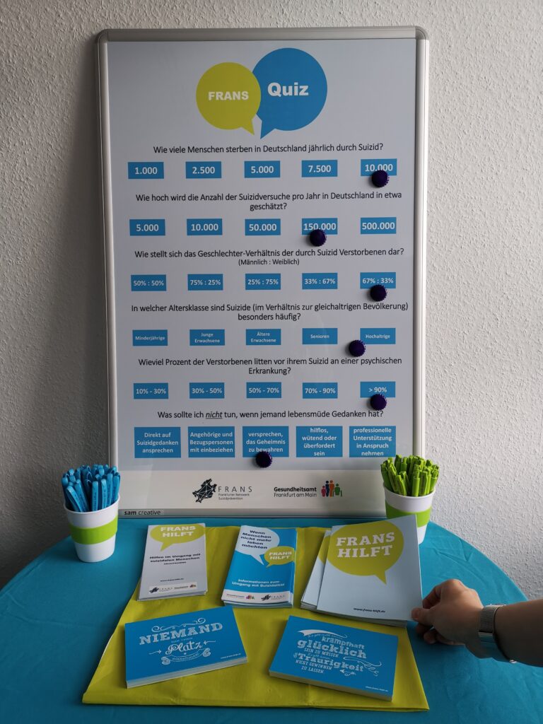 Kleiner, runder Tisch, auf dem das bunte FRANS Quiz steht und davor FRANS Stifte in Grün und Blau. Außerdem liegen ein paar Infomaterialien wie Flyer und Postkarten. Eine Hand greift nach dem FRANS hilft Flyer.