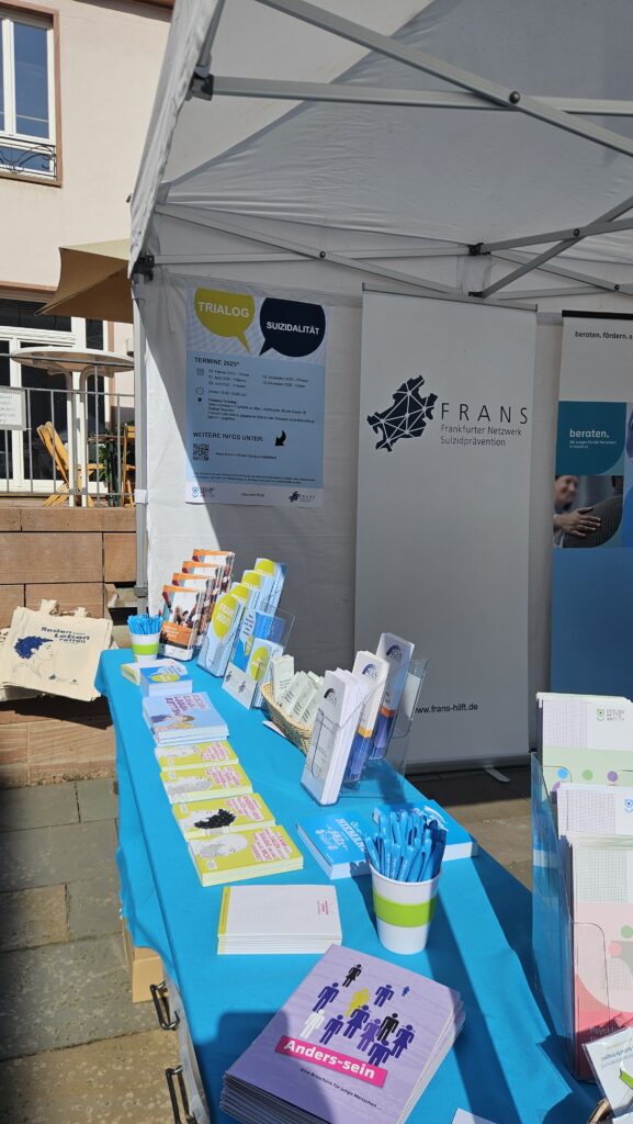 FRANS Infostand in einem weißen Pavillon. Tisch mit blauer Decke und darauf buntes Infomaterial wie Flyer und Postkarten. Im Hintergrund hängt ein Plakat zum Trialog Suizidalität und ein FRANS Roll-Up.