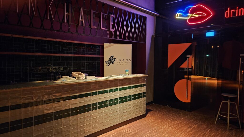 Theke im ZOOM Frankfurt. Auf der Theke liegen Infomaterialien von FRANS und dahinter steht ein weißes Roll-Up mit Logo. Drum herum kann man durch eine geöffnete Tür die Tanzfläche erahnen.